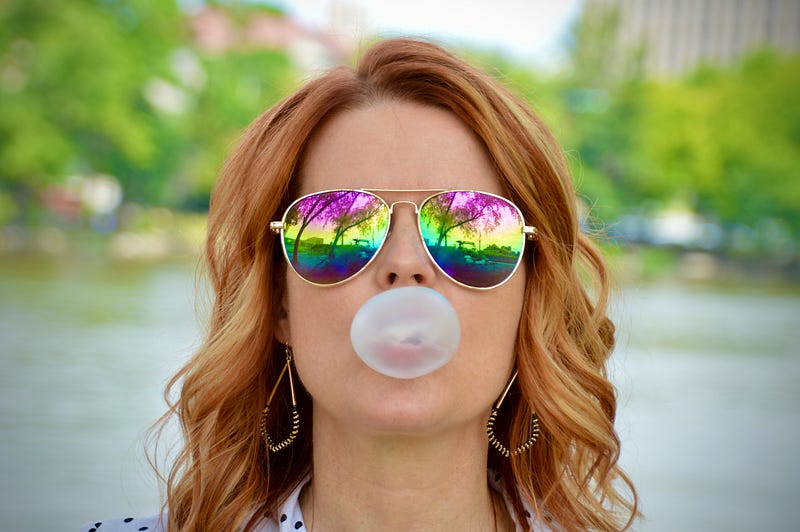 Playful woman with colorful glasses and a balloon in her mouth, expressing nonconformity and creative freedom.