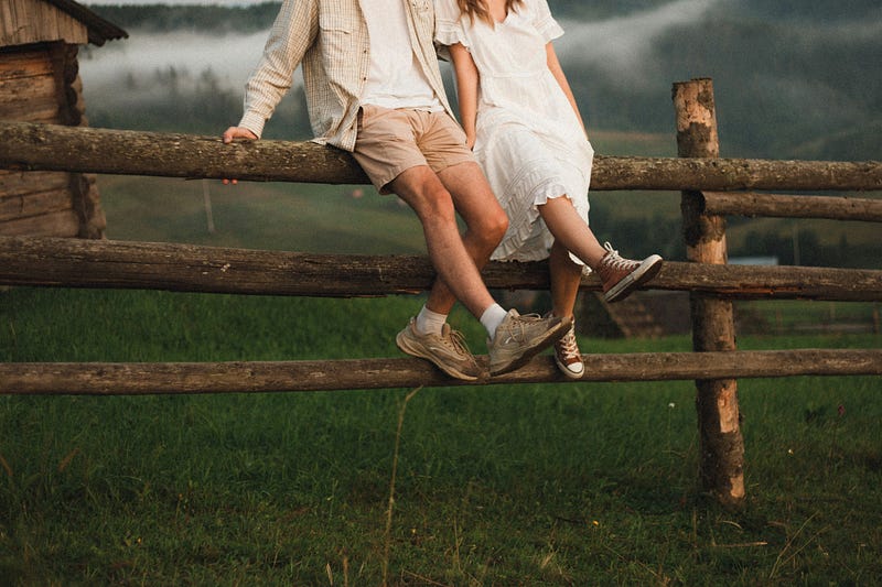 A couple sitting side by side on a wooden ranch fence, only their legs and shoes visible, symbolising quiet connection, intimacy, and unspoken understanding.
